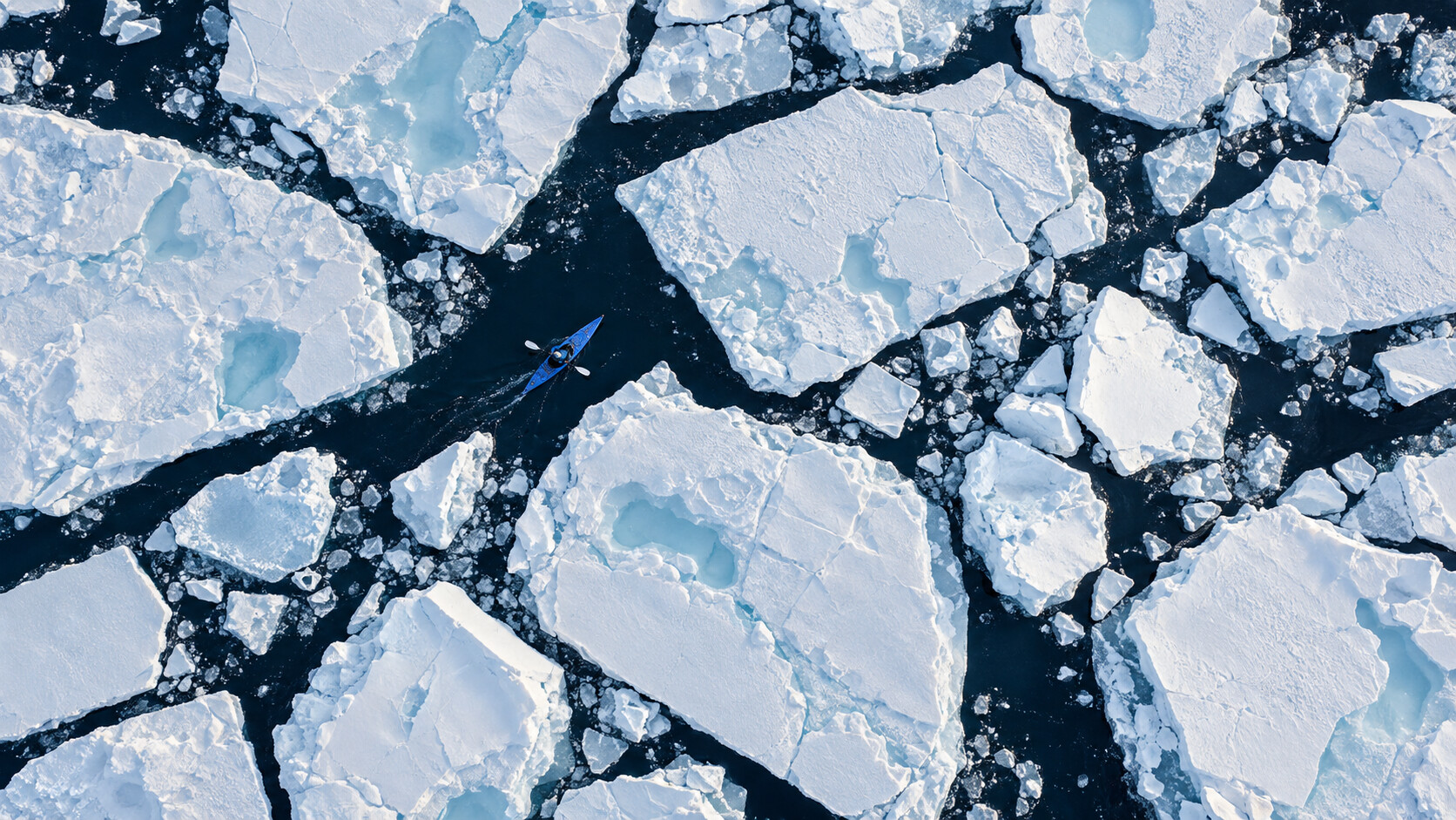 A lone kayaker paddles through channels between sheets of sea ice, seen from above.